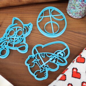 Op de afbeelding: Drie blauwe plastic koekjesvormen in de vorm van een videogamepersonage, een videogamepersonage met een hoed en een cirkel met een ontwerp erin.