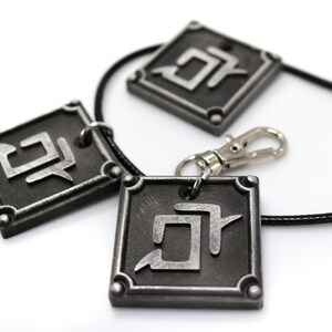 K&ouml;nnte beinhalten: Drei silberfarbene Anh&auml;nger mit einem schwarzen Band und einem silbernen Verschluss. Jeder Anh&auml;nger hat eine quadratische Form mit einem stilisierten Symbol im Inneren. Das Symbol ist ein stilisierter Buchstabe "D" mit einem diagonalen Strich durch ihn.