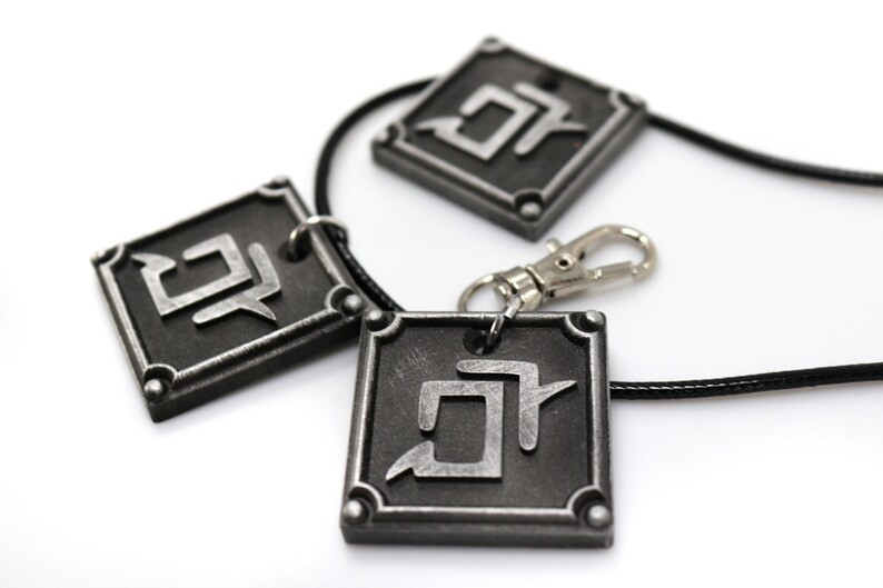 K&ouml;nnte beinhalten: Drei silberfarbene Anh&auml;nger mit einem schwarzen Band und einem silbernen Verschluss. Jeder Anh&auml;nger hat eine quadratische Form mit einem stilisierten Symbol im Inneren. Das Symbol ist ein stilisierter Buchstabe "D" mit einem diagonalen Strich durch ihn.