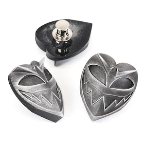 May include: Three silver-colored metal pins with a heart-shaped design. The pins feature a stylized, menacing face with sharp teeth and eyes. One pin is a black spade shape with a silver-colored pin back.