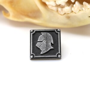 May include: A silver metal pin with a square design. The pin features a raised image of a knight's helmet in a silver finish.