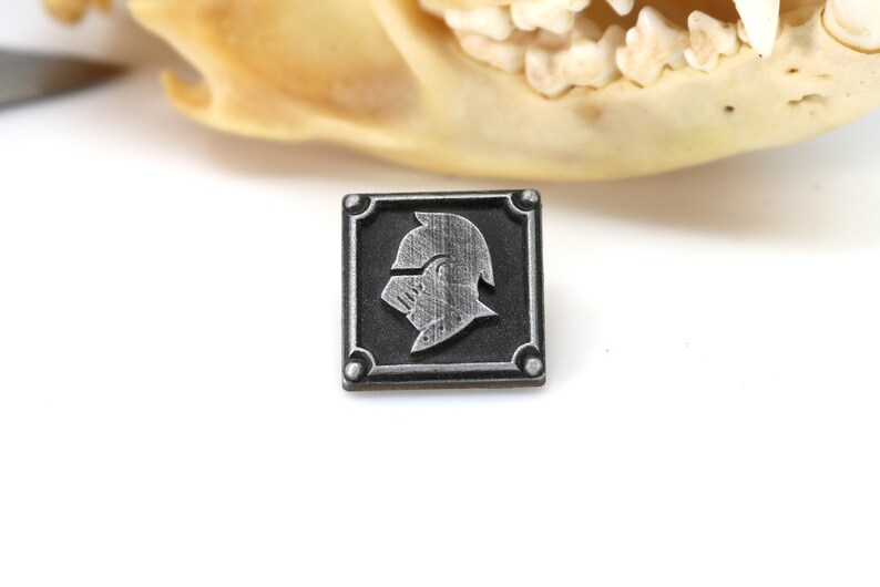 May include: A silver metal pin with a square design. The pin features a raised image of a knight's helmet in a silver finish.