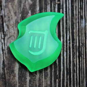 May include: A translucent green shield-shaped pendant with a lighter green interior. The interior features a stylized design of three vertical lines and a curved line. The pendant is set against a weathered wooden background.