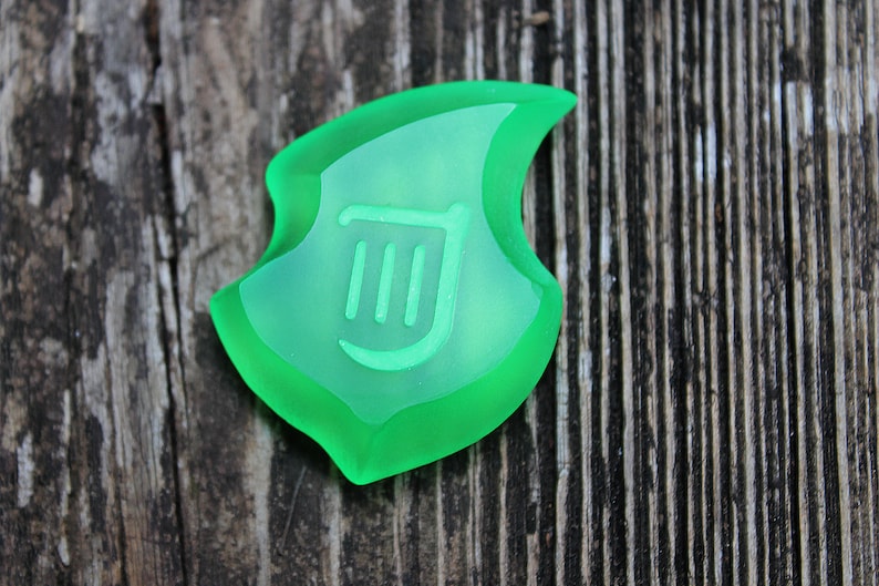 May include: A translucent green shield-shaped pendant with a lighter green interior. The interior features a stylized design of three vertical lines and a curved line. The pendant is set against a weathered wooden background.