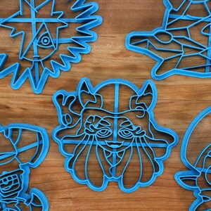 May include: Five blue plastic cookie cutters shaped like cartoon characters. The cutters are on a wooden surface.
