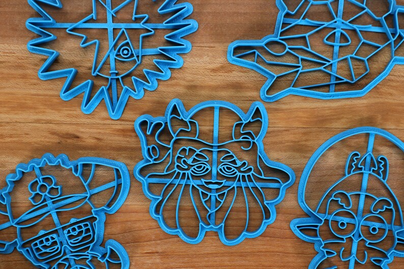May include: Five blue plastic cookie cutters shaped like cartoon characters. The cutters are on a wooden surface.