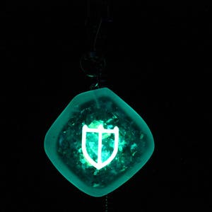 May include: A teal blue pendant with a white shield design. The pendant is hanging from a black cord.