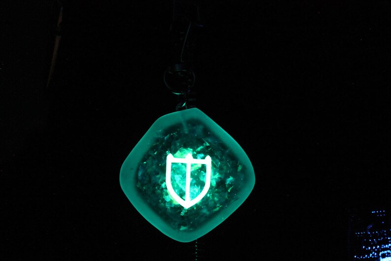 May include: A teal blue pendant with a white shield design. The pendant is hanging from a black cord.