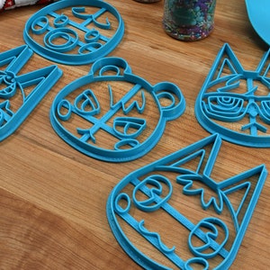 May include: Five blue plastic cookie cutters shaped like animal faces. The cutters are on a wooden surface. The animal faces include a cat, a bear, a fox, and two different rabbits.