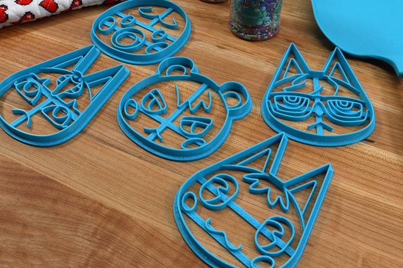 May include: Five blue plastic cookie cutters shaped like animal faces. The cutters are on a wooden surface. The animal faces include a cat, a bear, a fox, and two different rabbits.
