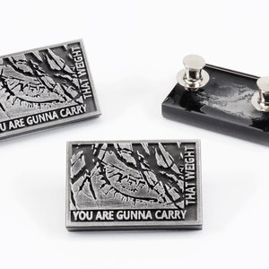 May include: Two silver pins with a cracked, textured design and the text "You are gunna carry that weight".