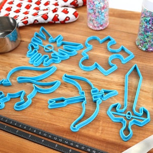 May include: A set of blue plastic cookie cutters shaped like a sword, bow and arrow, a puzzle piece, a winged creature, and a dog. The cutters are on a wooden surface with a rolling pin, a measuring cup, a spatula, and two jars of sprinkles.