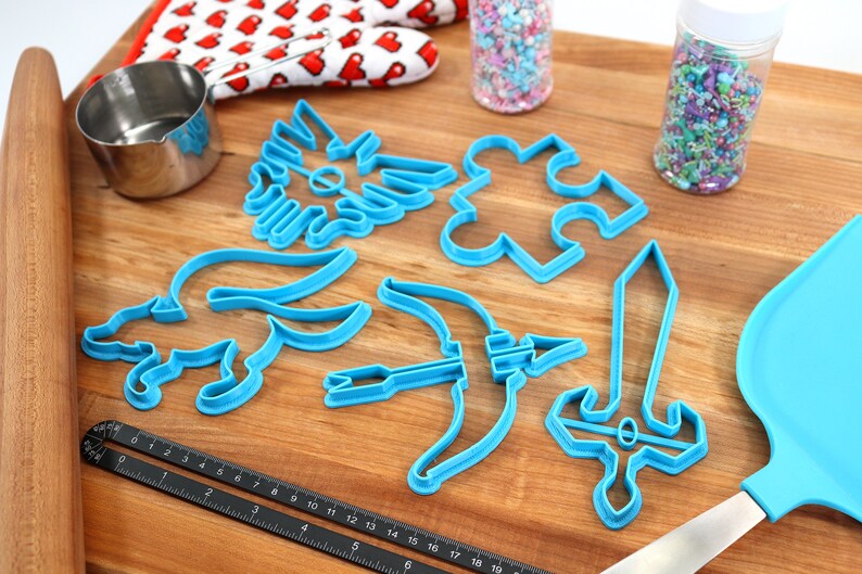 May include: A set of blue plastic cookie cutters shaped like a sword, bow and arrow, a puzzle piece, a winged creature, and a dog. The cutters are on a wooden surface with a rolling pin, a measuring cup, a spatula, and two jars of sprinkles.