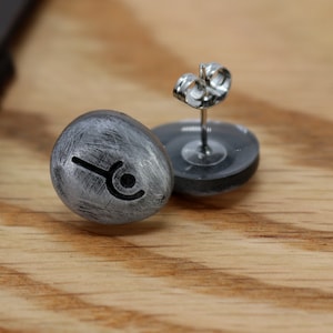 May include: A pair of silver stud earrings with a black symbol on each. The earrings are on a wooden surface.