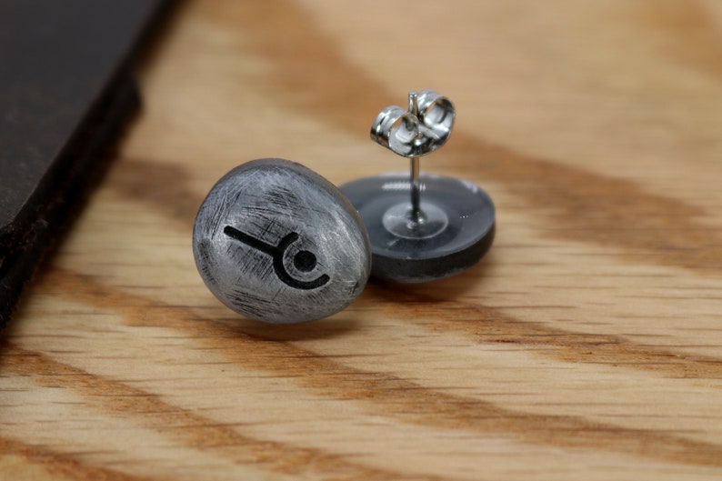 May include: A pair of silver stud earrings with a black symbol on each. The earrings are on a wooden surface.
