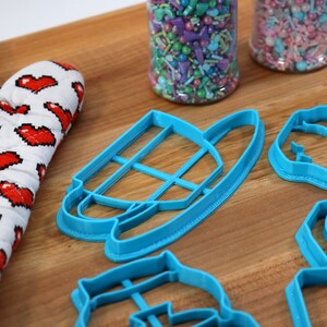 May include: Four blue plastic cookie cutters on a wooden surface. The cutters are shaped like a teacup, a house, a flower, and a heart.