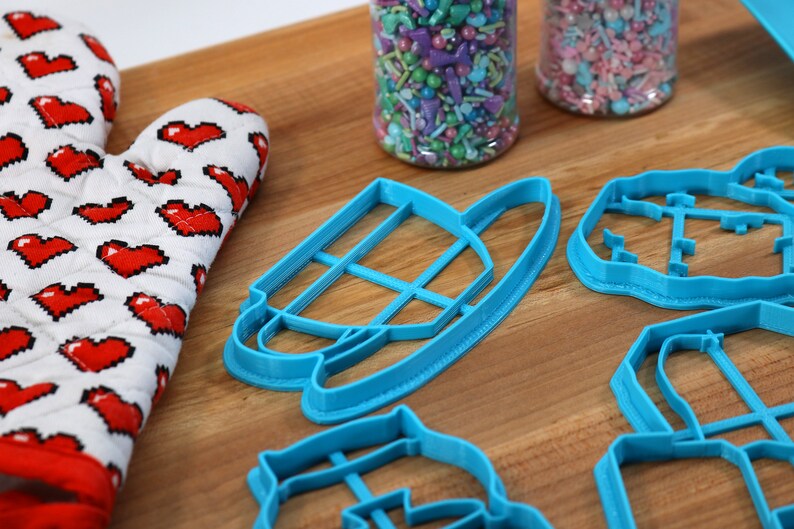 May include: Four blue plastic cookie cutters on a wooden surface. The cutters are shaped like a teacup, a house, a flower, and a heart.