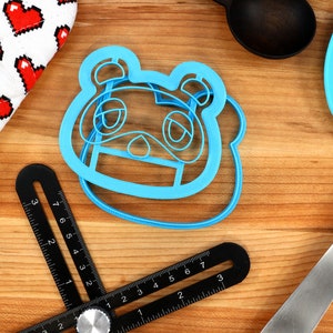 Może przedstawiać: Niebieska plastikowa foremka do ciasteczek w kształcie postaci "Tom Nook" z gry wideo Animal Crossing. Foremka leży na drewnianej powierzchni.