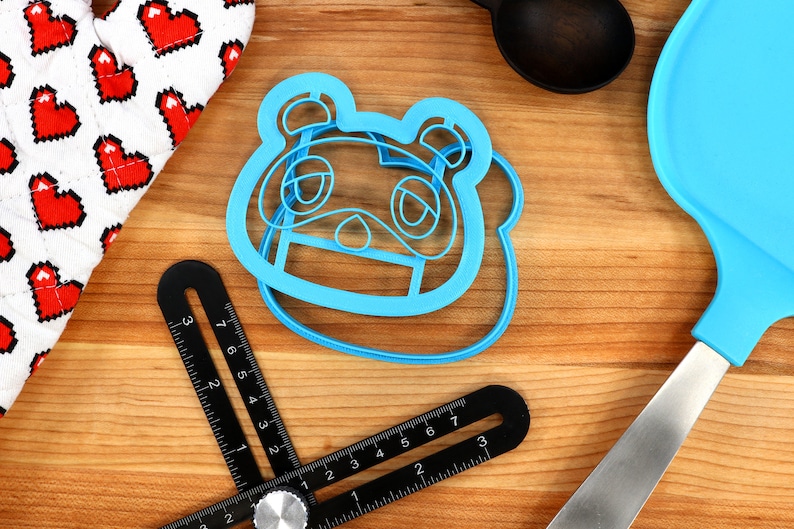 Może przedstawiać: Niebieska plastikowa foremka do ciasteczek w kształcie postaci "Tom Nook" z gry wideo Animal Crossing. Foremka leży na drewnianej powierzchni.