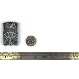 May include: A silver metal charm featuring a stylised eye design and geometric patterns. The charm is approximately 5 cm tall and is next to a US dime for scale.