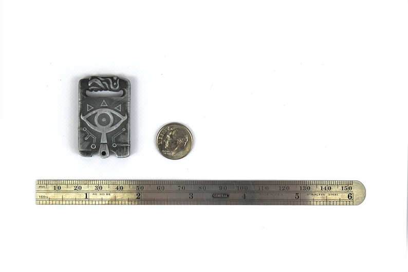 May include: A silver metal charm featuring a stylised eye design and geometric patterns. The charm is approximately 5 cm tall and is next to a US dime for scale.