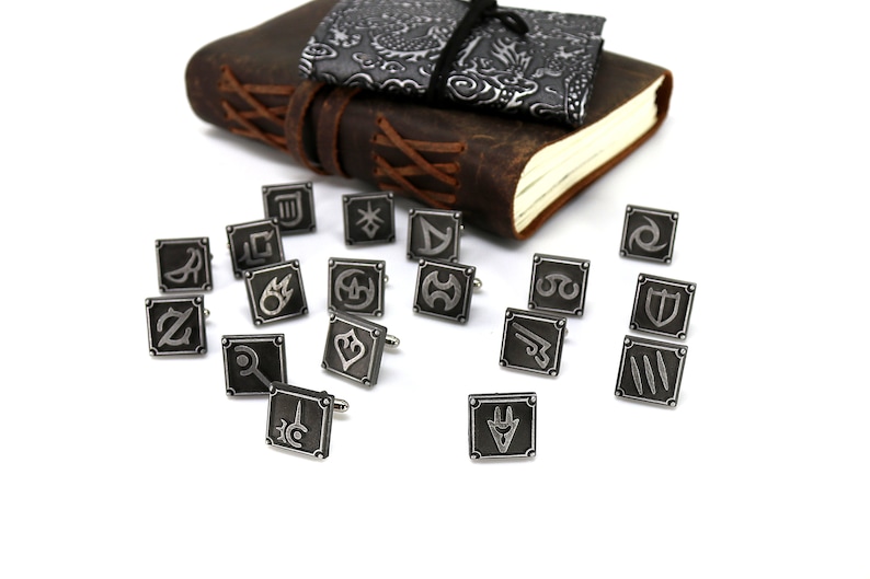May include: A set of 16 silver cufflinks featuring various symbols, including a sword, a shield, and a crown. The cufflinks are arranged on a white surface.