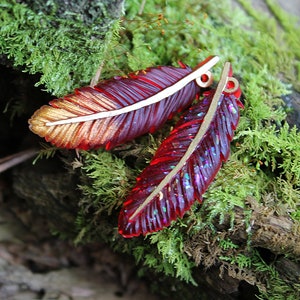 May include: Two red and gold resin feather charms with glitter accents. The feathers are on a bed of green moss.