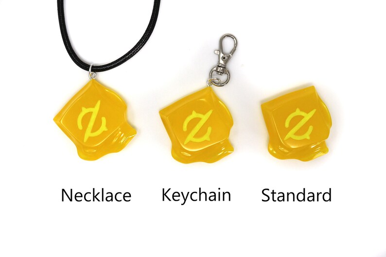 以下が含まれることがあります： スタイリッシュな「Z」デザインのイエローペンダント。ネックレス、キーチェーン、標準チャームとして利用可能。ネックレスは黒いコード付き。キーチェーンはシルバーの留め金付き。「Necklace」、「Keychain」、「Standard」の文字が各アイテムの下にあります。