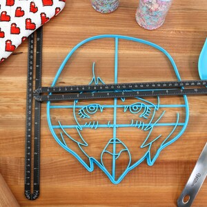 May include: A blue plastic cookie cutter shaped like a cartoon face with large eyes and a mouth. The cutter is on a wooden surface with a ruler and a spatula.
