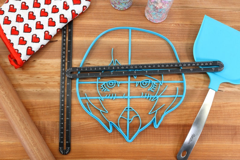 May include: A blue plastic cookie cutter shaped like a cartoon face with large eyes and a mouth. The cutter is on a wooden surface with a ruler and a spatula.