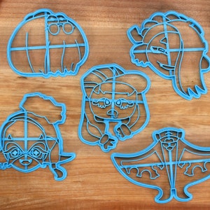 May include: Five blue plastic cookie cutters shaped like cartoon characters. The cutters are on a wooden surface.
