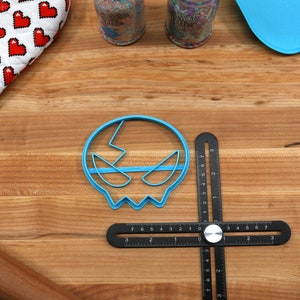 May include: A blue plastic skull-shaped cookie cutter on a wooden surface with a black ruler and a rolling pin.
