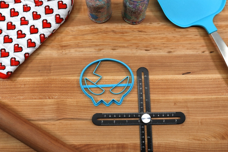 May include: A blue plastic skull-shaped cookie cutter on a wooden surface with a black ruler and a rolling pin.