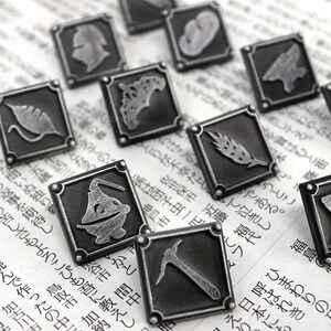 May include: A set of ten silver-toned metal pins with different designs. Each pin is square-shaped and features a different design, including a leaf, a wheat stalk, a pickaxe, and a creature.