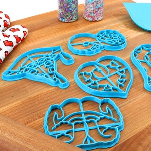 May include: A set of five blue plastic cookie cutters in various shapes, including a heart, a donut, a flower, and two abstract designs. The cutters are on a wooden surface with a rolling pin and a spatula.
