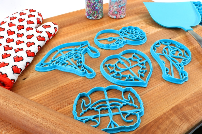 May include: A set of five blue plastic cookie cutters in various shapes, including a heart, a donut, a flower, and two abstract designs. The cutters are on a wooden surface with a rolling pin and a spatula.