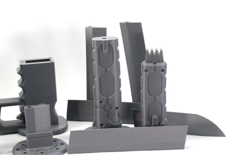 Puede incluir: Un conjunto de piezas grises impresas en 3D para una pistola de modelo. Las piezas incluyen un ca&ntilde;&oacute;n, una empu&ntilde;adura, un cargador y una mira.