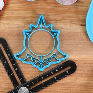 May include: A blue plastic cookie cutter shaped like a stylized sun with a circle in the center. The cookie cutter is on a wooden surface.