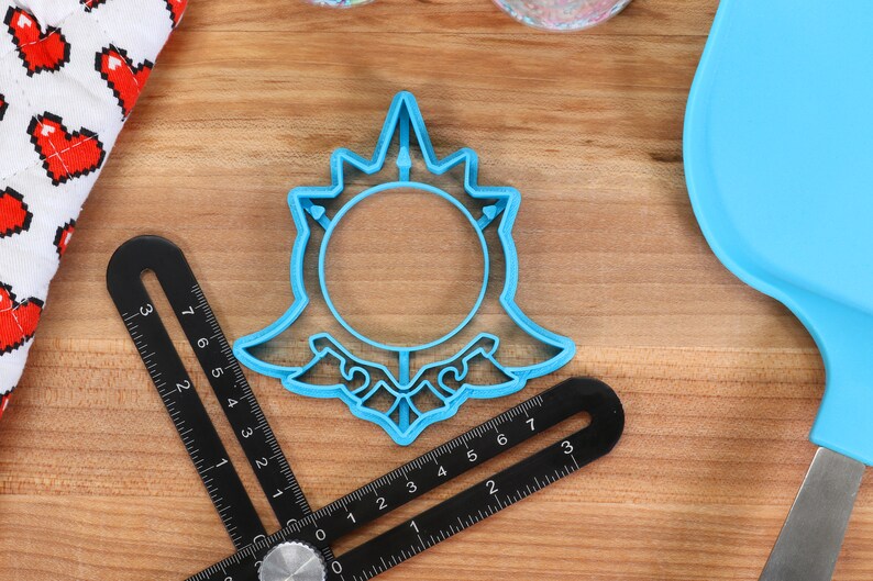 May include: A blue plastic cookie cutter shaped like a stylized sun with a circle in the center. The cookie cutter is on a wooden surface.