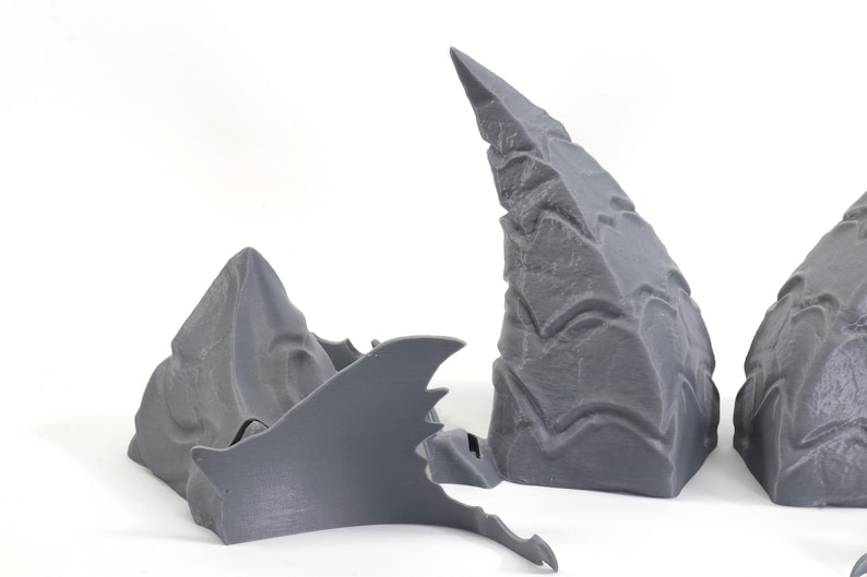 Op de afbeelding: Drie grijze 3D-geprinte onderdelen, twee zijn hoog en puntig, en &eacute;&eacute;n is plat en vleugelvormig.