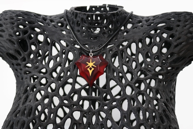 May include: A red heart-shaped pendant necklace with a gold symbol inside. The necklace is on a black mannequin bust.