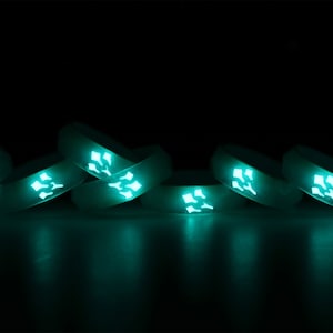 May include: A group of eight glow-in-the-dark silicone rings stacked on top of each other. The rings are a light teal color and have a geometric design.