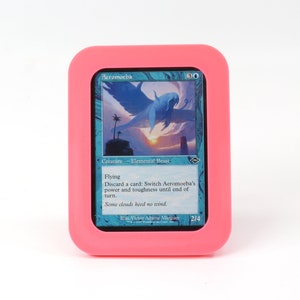 Op de afbeelding: Een roze kaarthouder met een blauwe en witte Magic: The Gathering kaart erin. De kaart heet "Aeromoeba" en is een wezen - elementair beest met de mogelijkheid om te vliegen. De tekst op de kaart luidt: "Vliegen. Een kaart afleggen: Wissel de kracht en taaiheid van Aeromoeba om tot het einde van de beurt. Sommige wolken houden geen rekening met de wind."