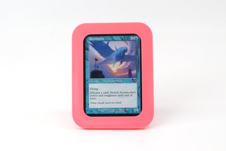Op de afbeelding: Een roze kaarthouder met een blauwe en witte Magic: The Gathering kaart erin. De kaart heet "Aeromoeba" en is een wezen - elementair beest met de mogelijkheid om te vliegen. De tekst op de kaart luidt: "Vliegen. Een kaart afleggen: Wissel de kracht en taaiheid van Aeromoeba om tot het einde van de beurt. Sommige wolken houden geen rekening met de wind."