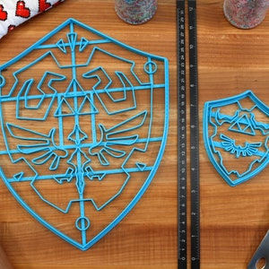 May include: Two blue plastic cookie cutters shaped like the Triforce shield from the Legend of Zelda video game series. The larger cutter has a detailed design with a grid pattern, while the smaller cutter has a more simplified design.