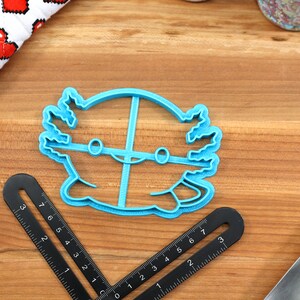 May include: A blue plastic cookie cutter shaped like an axolotl, a type of salamander. The cutter has a simple design with two eyes and a smile.