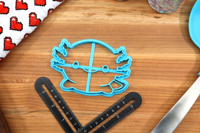 May include: A blue plastic cookie cutter shaped like an axolotl, a type of salamander. The cutter has a simple design with two eyes and a smile.
