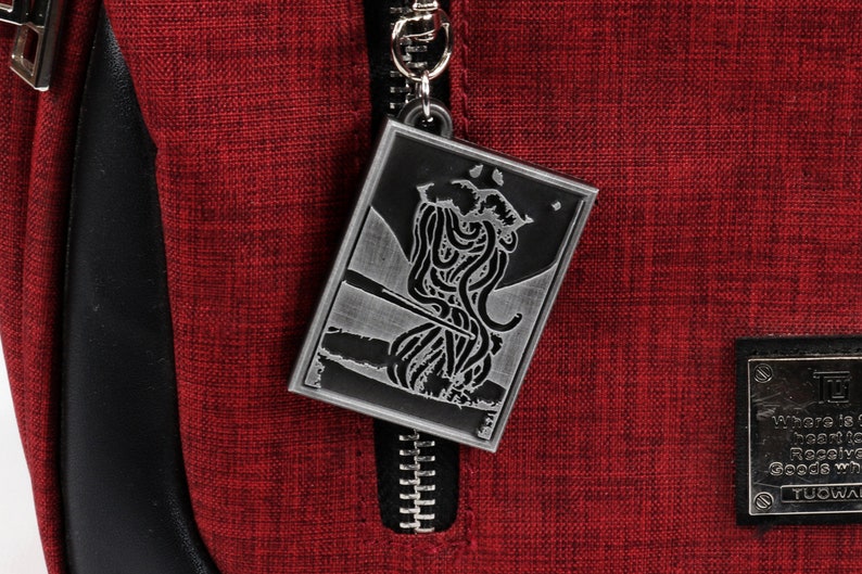 May include: A silver zipper pull with a detailed design of a creature with tentacles. The zipper pull is attached to a red fabric bag.