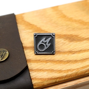 Puede incluir: Un pin de metal plateado con fondo negro y un dise&ntilde;o de llama estilizado. El pin est&aacute; sobre una superficie de madera.