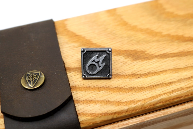 Puede incluir: Un pin de metal plateado con fondo negro y un dise&ntilde;o de llama estilizado. El pin est&aacute; sobre una superficie de madera.
